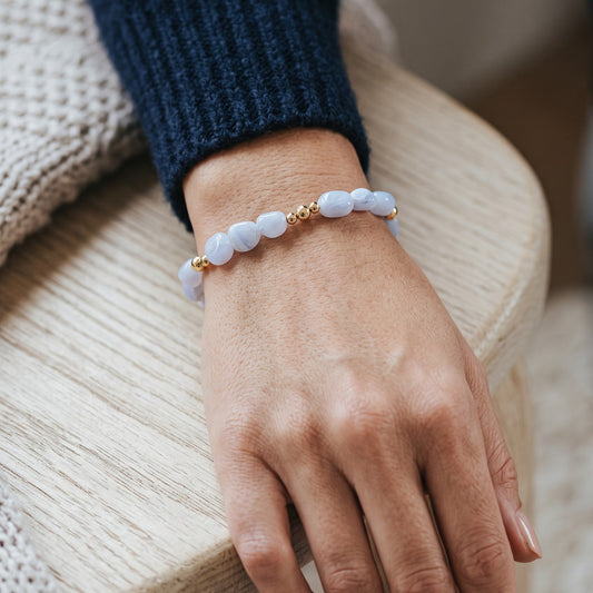A close-up of a person's hand wearing a delicate beaded bracelet. The bracelet features Blue Lace Agate nugget beads separated by small gold spacer beads, worn over a dark navy blue knit sleeve.