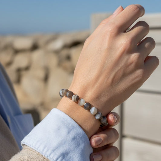 A close-up of a person's wrist wearing a beaded bracelet made of polished brown, gray, and white agate-like stones, with a light blue shirt cuff visible against a blurred outdoor background.