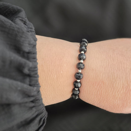 A close-up of a person's wrist wearing a beaded bracelet made of faceted Black Tourmaline beads separated by small silver spacer beads, with a dark gray sleeve in the foreground.