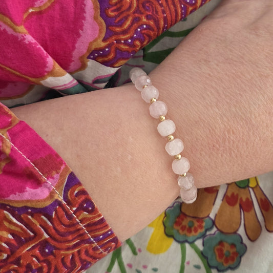 A close-up of a person's wrist wearing a beaded bracelet made of pale pink Rose Quartz beads separated by small gold spacer beads, against a colorful floral-patterned garment.