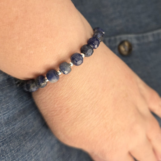 A close-up of a beaded bracelet worn on a person's wrist, featuring faceted blue Sodalite beads separated by small silver spacer beads, set against a background of blue denim.