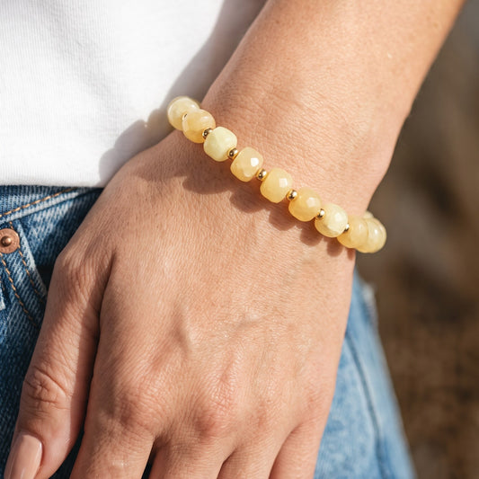 A close-up of a person's wrist wearing a beaded stretch bracelet made of Yellow Calcite faceted beads separated by small gold spacer beads, worn over a white shirt and blue jeans.