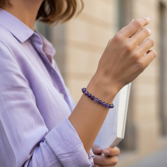 A person wearing a purple beaded bracelet on their wrist, wearing a light purple button-up shirt, with a blurred outdoor background.