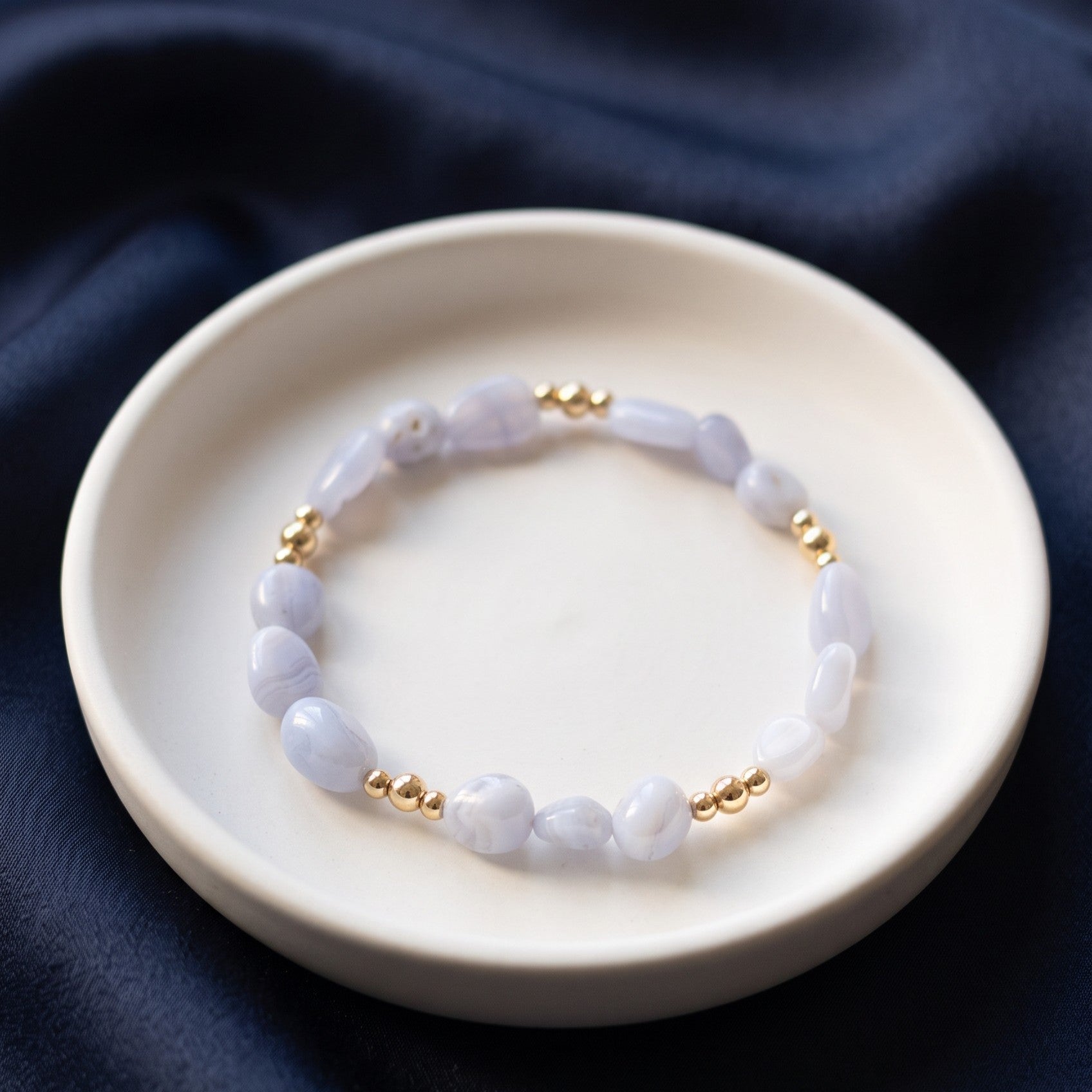 A beaded bracelet with Blue Lace Agate nugget beads and small gold spacer beads, displayed on a white ceramic dish over dark navy fabric.