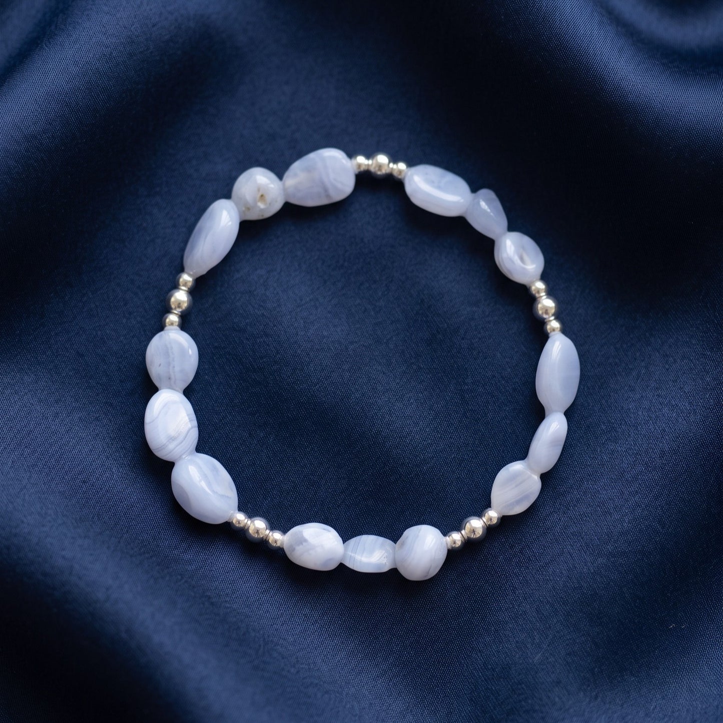 A beaded stretch bracelet featuring Blue Lace Agate nugget beads and small metallic silver spacer beads, displayed on a dark navy blue satin fabric background.