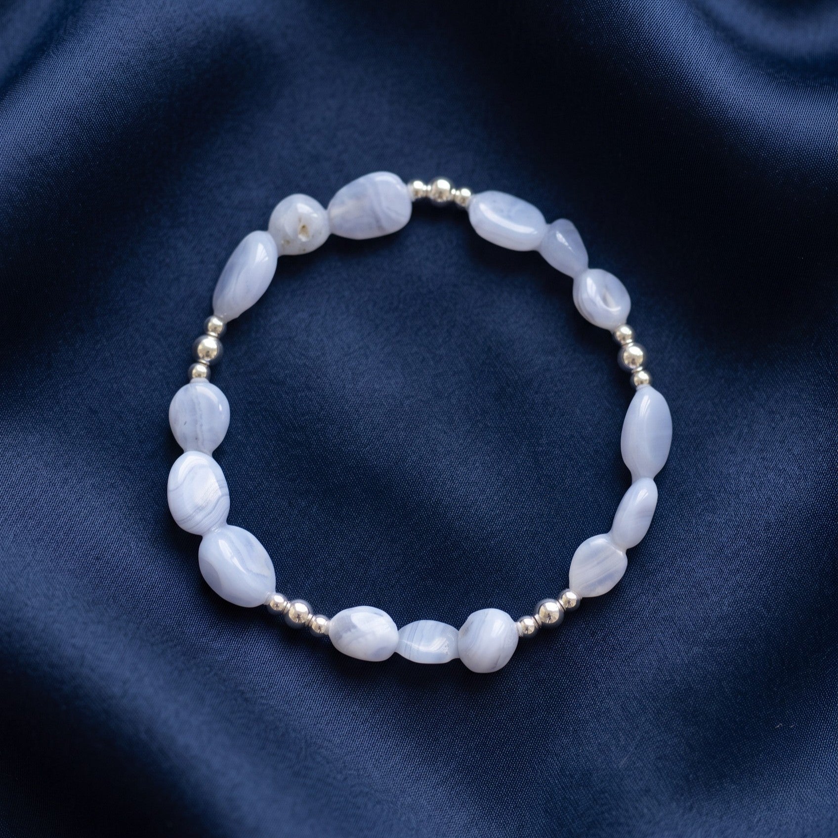 A beaded stretch bracelet featuring Blue Lace Agate nugget beads and small metallic silver spacer beads, displayed on a dark navy blue satin fabric background.