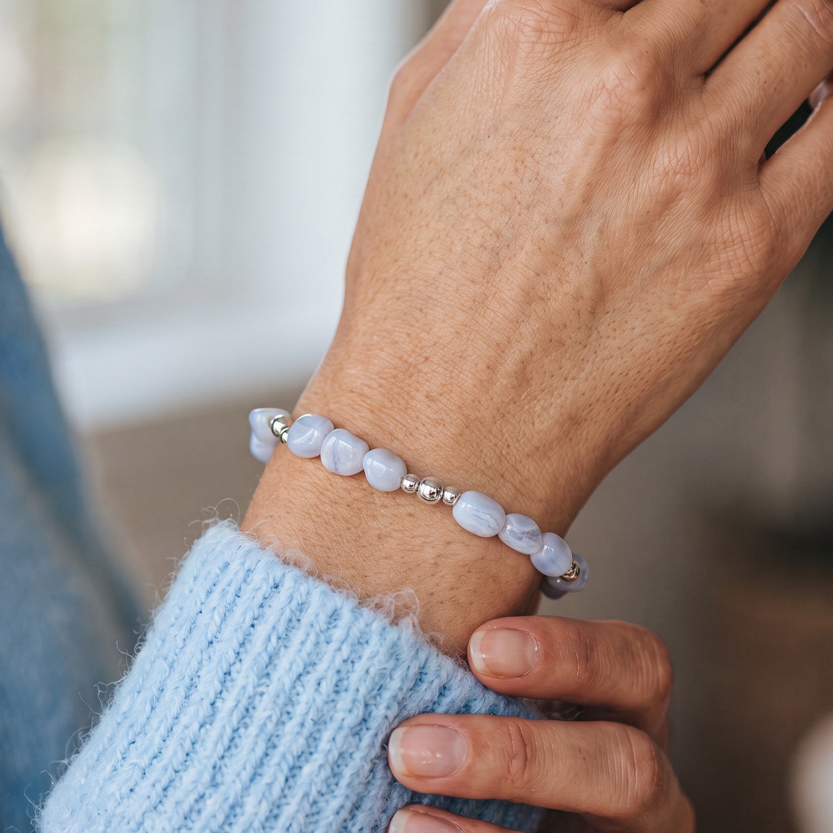 A close-up of a person's wrist wearing a beaded bracelet made of Blue Lace Agate nugget beads separated by small silver spacer beads, paired with a light blue knit sweater.