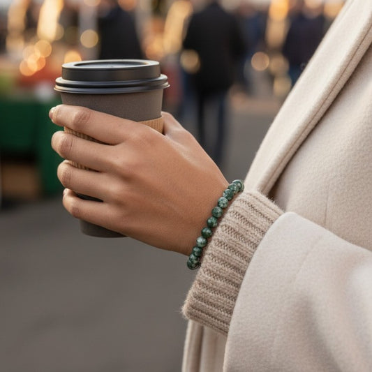 A close-up of a person's hand holding a takeaway coffee cup, wearing a green beaded bracelet on the wrist. The person is dressed in a beige coat and ribbed beige sweater, with a blurred outdoor market background.