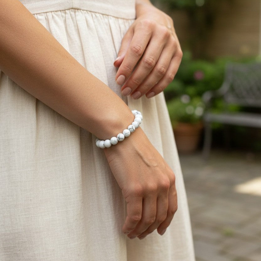 A close-up of a woman wearing a white beaded bracelet on her wrist, dressed in a light beige linen dress, with a softly blurred outdoor garden background.
