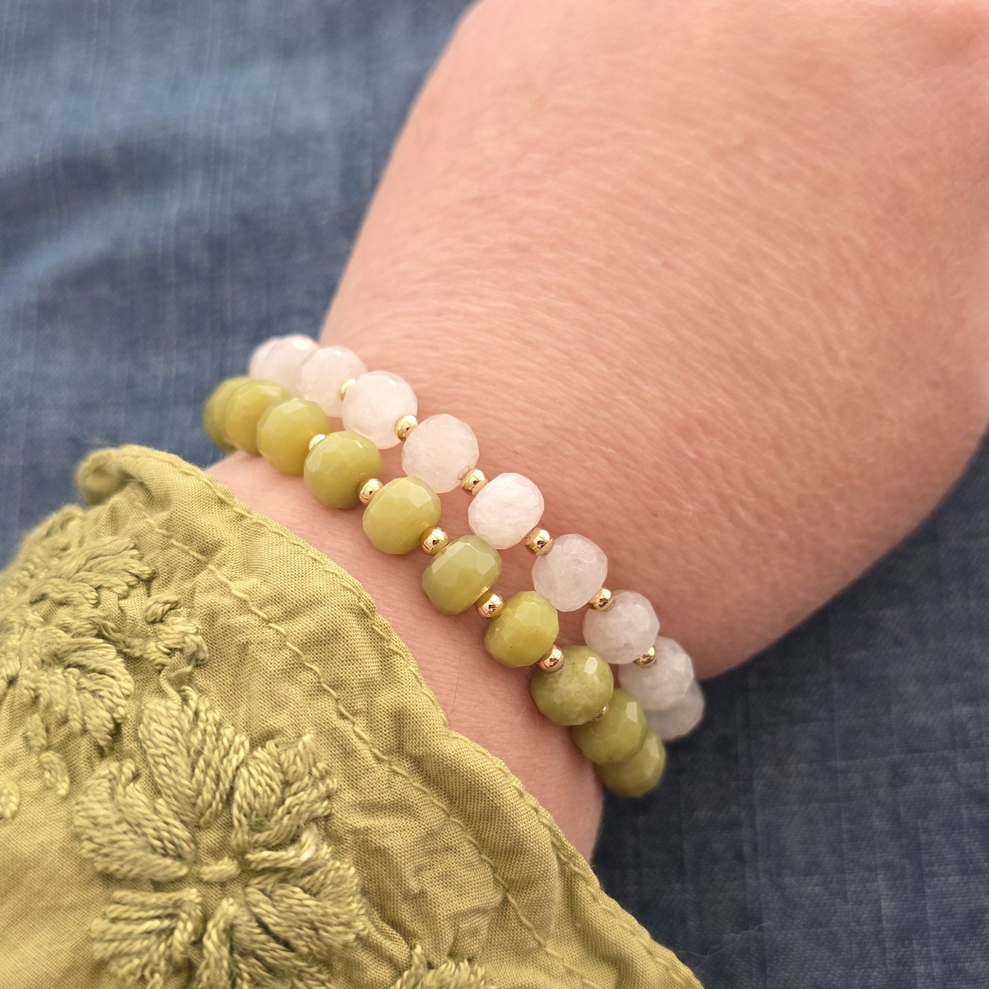 A close-up of a person's wrist wearing two beaded stretch bracelets. One bracelet features faceted green Serpentine beads with small gold spacer beads, and the other features faceted white Moonstone beads with gold spacer beads.