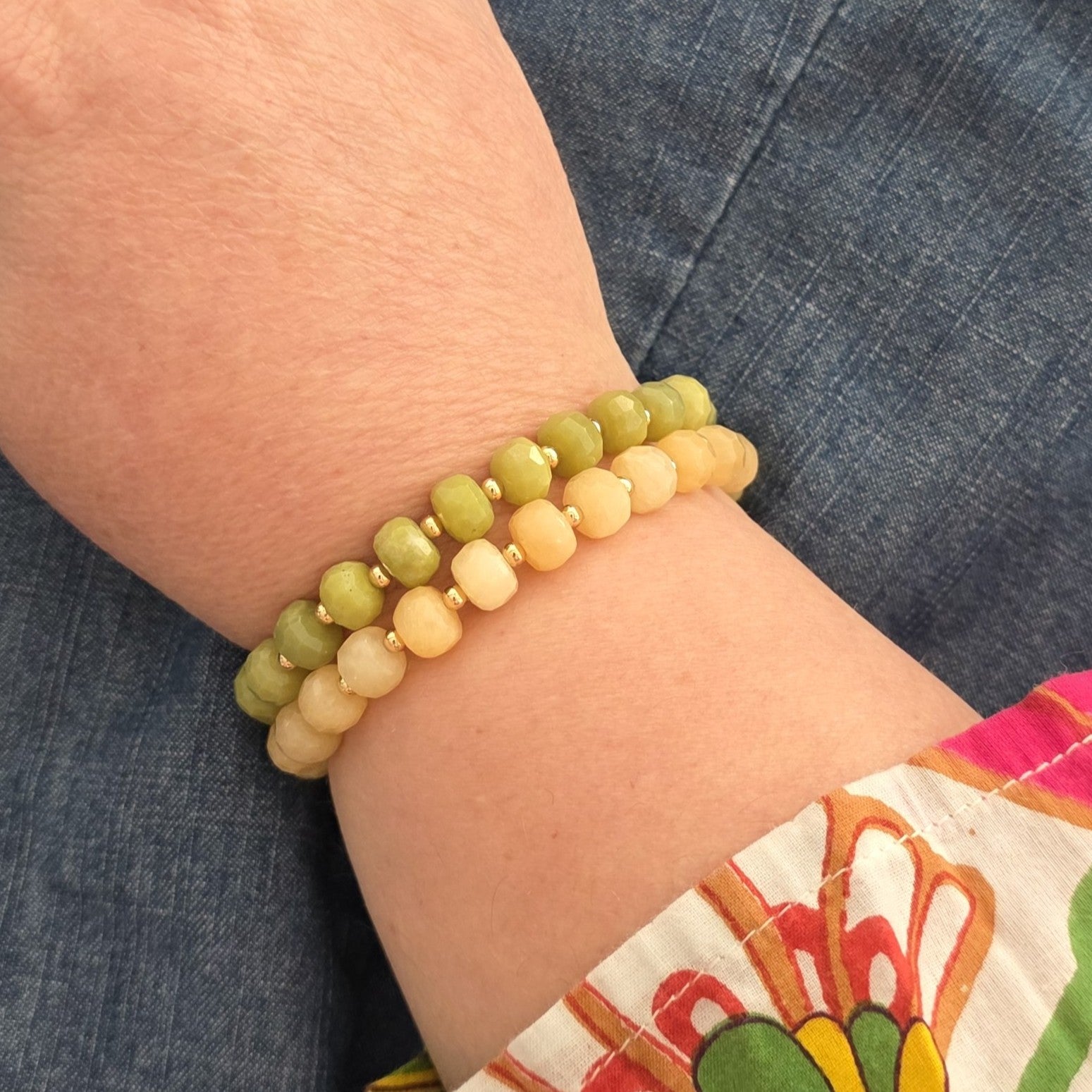 Two beaded stretch bracelets on a person's wrist, one featuring green Serpentine faceted beads separated by small gold spacer beads and one featuring Yellow Calcite faceted beads separated by small gold space beads, shown against blue denim clothing.