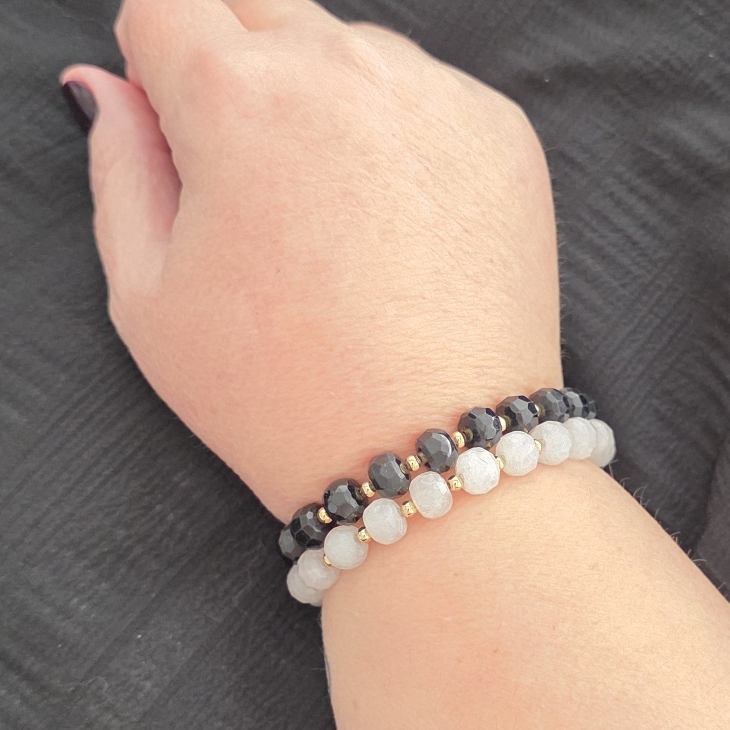 A close-up of a person's wrist wearing two beaded stretch bracelets. One bracelet features Black Tourmaline faceted beads with small gold spacer rings, and the other features white Moonstone round beads with small gold spacer rings.