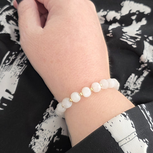 A close-up of a person's wrist wearing a beaded bracelet made of white round  Moonstone beads separated by small gold spacer beads, resting against a black and white patterned garment.