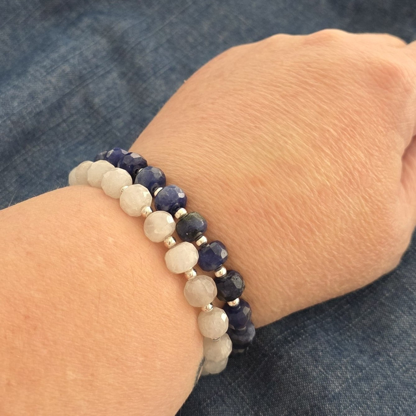 Two beaded stretch bracelets worn on a person's wrist. One bracelet features faceted blue Sodalite and silver spacer beads, while the other features faceted white Moonstone beads with small silver spacers.