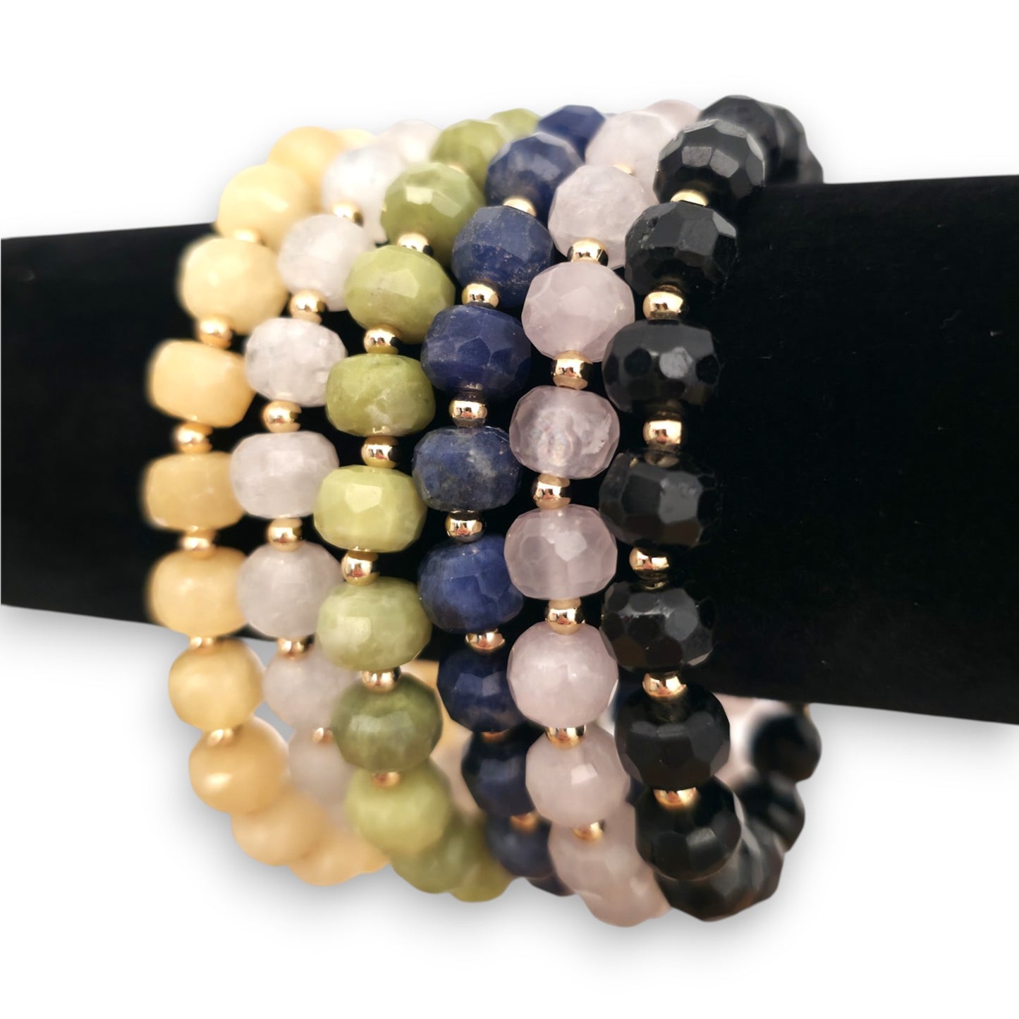 A set of beaded stretch bracelets displayed on a black stand. The bracelets feature faceted beads in various colors including Yellow Calcite, White Moonstone, green Serpentine, blue Sodalite, pink Rose Quartz and Black Tourmaline, separated by small gold spacer beads.