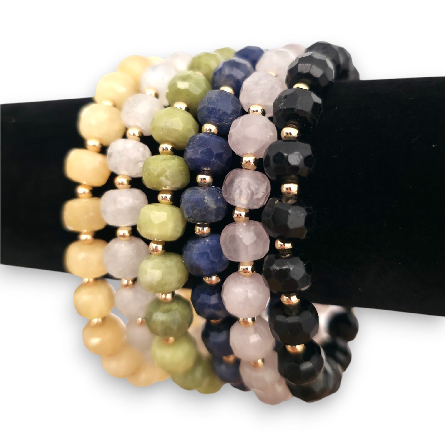 A set of beaded stretch bracelets displayed on a black stand. The bracelets feature faceted beads in various colors including Yellow Calcite, White Moonstone, green Serpentine, blue Sodalite, pink Rose Quartz and Black Tourmaline, separated by small gold spacer beads.