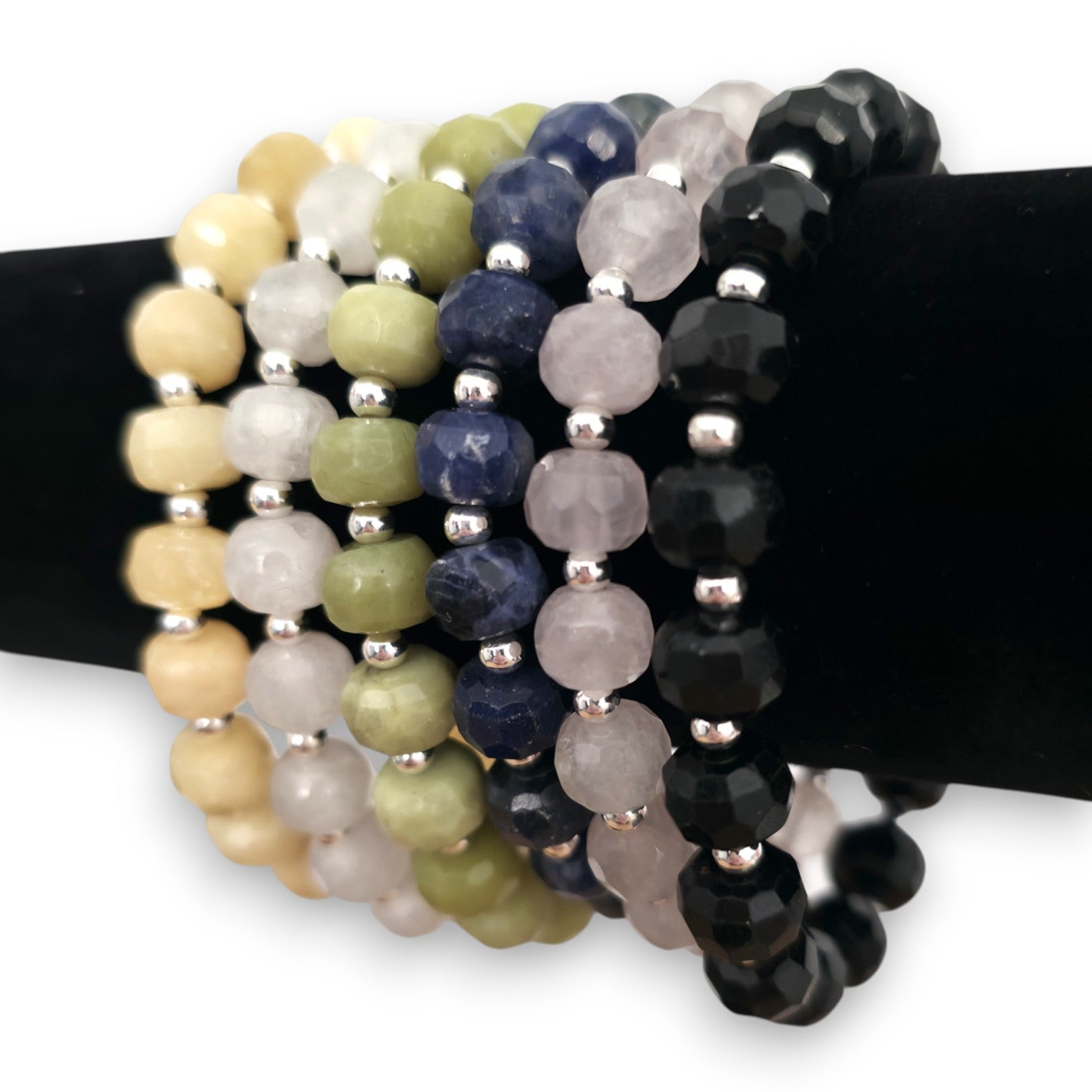 A set of beaded stretch bracelets displayed on a black stand. The bracelets feature faceted beads in various colors including Yellow Calcite, White Moonstone, green Serpentine, blue Sodalite, pink Rose Quartz and Black Tourmaline, separated by small silver spacer beads.
